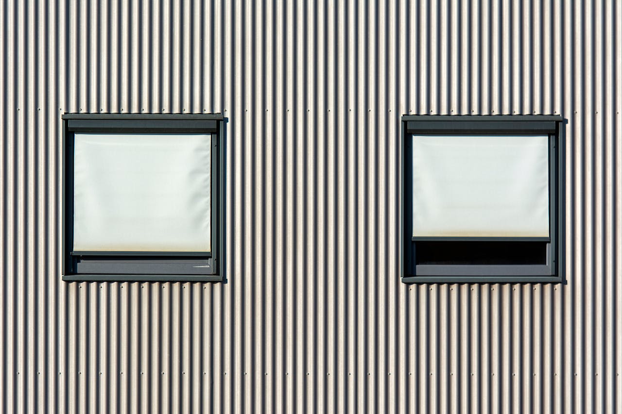 Minimalist architectural facade featuring two windows with closed blinds and corrugated metal siding.